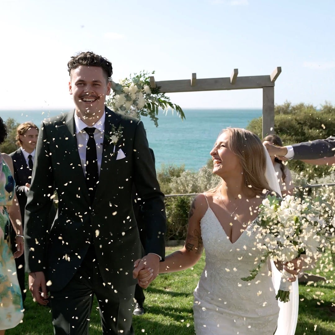 A bride and groom laughing hand in hand as guests shower them with petals by the sea